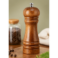 A wooden pepper mill grinder with a polished brown wood finish and a silver metal top, standing on a wooden cutting board with a blurred jar of peppercorns in the background.